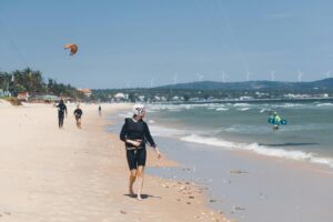 Kitesurfers walk and surf on a sunny beach with wind turbines in the background, capturing a vibrant seaside activity scene.